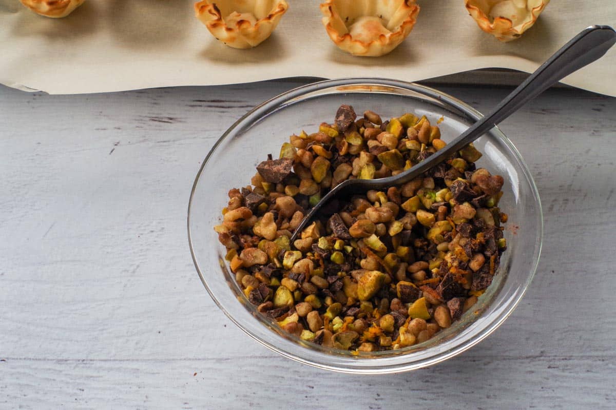 The chopped pistachios, walnuts, chopped chocolate, cinnamon, and orange zest stirred together in a small glass bowl with a spoon.