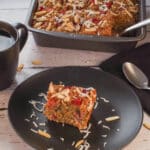 A slice of Baked Rhubarb Oatmeal on a black plate, with a cup of coffee and a pan with more Rhubarb Baked Oatmeal in the background.