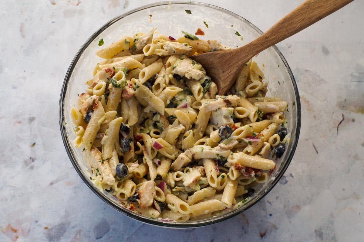 Tzatziki pasta mixed with all other ingredients in a glass bowl with a wooden spoon.