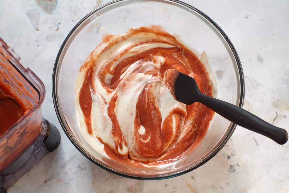 Rhubarb mixture folded into yogurt with black spatula in glass bowl.