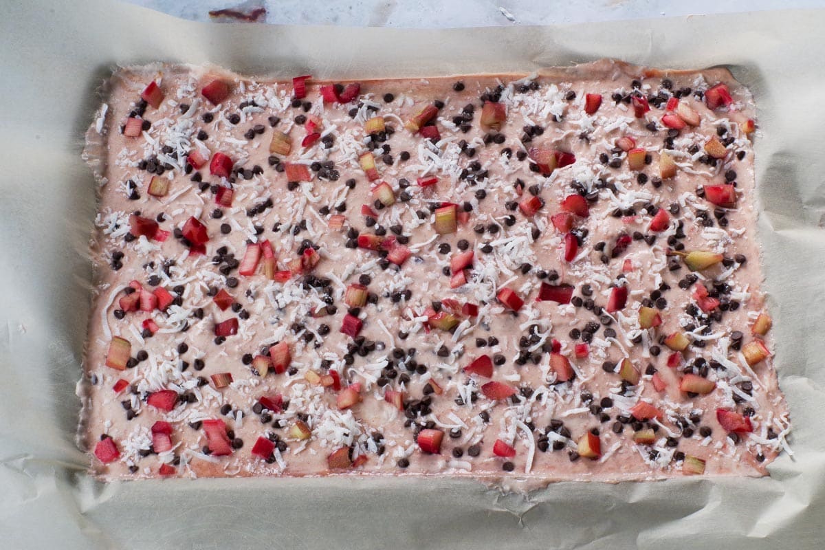 Rhubarb Yogurt spread on a parchment lined baking sheet with coconut, chocolate chips and rhubarb sprinkled on top.