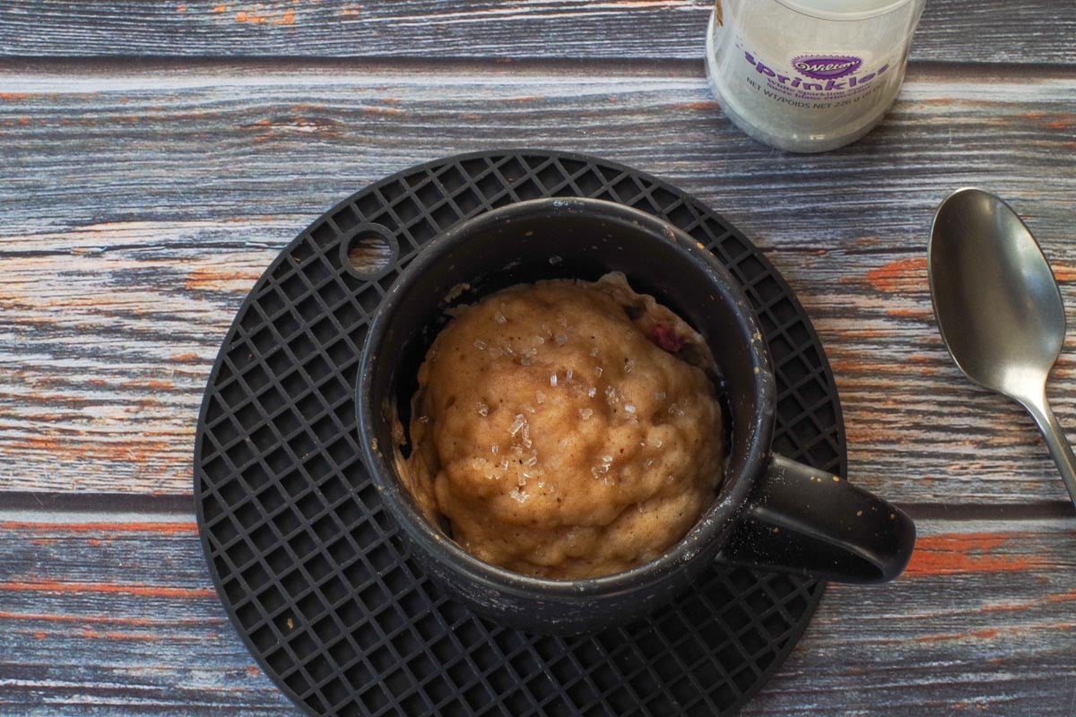 Baked Rhubarb Microwave Muffin in a mug, on a black trivet with sparkling sugar in the background.