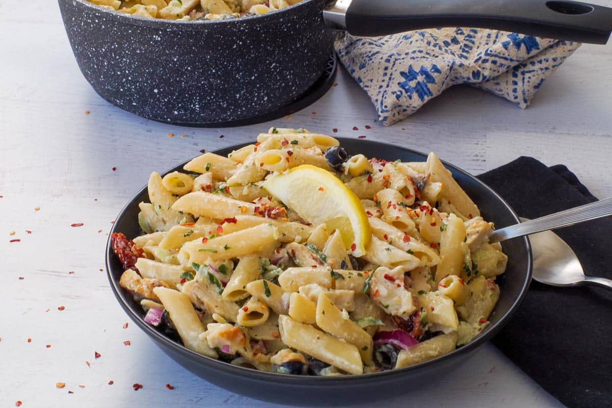 Tzatziki Pasta in a black bowl with a lemon wedg eon top and a black napkin beside the bowl and a pot in the background.