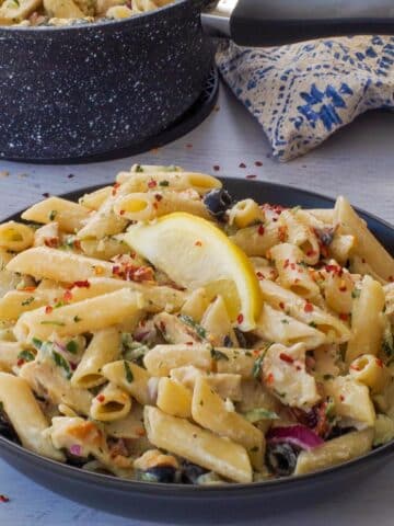 Tzatziki Pasta in a black bowl with a lemon wedg eon top and a black napkin beside the bowl and a pot in the background.