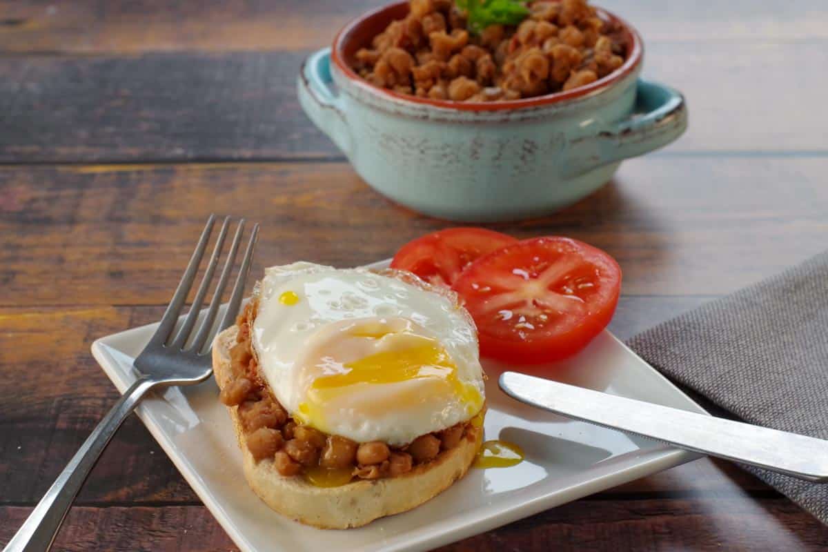 Baked Bean and Jalapeno Aoili on toast with an egg on top and a tomato on the side of a white plate and maple baked beans in the background.