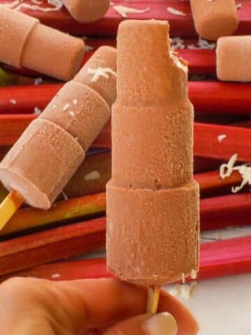Chocolate Rhubarb Popsicle (with a bite taken out at the top) being held up in front of a pile of rhubarb with more popsicles on top .