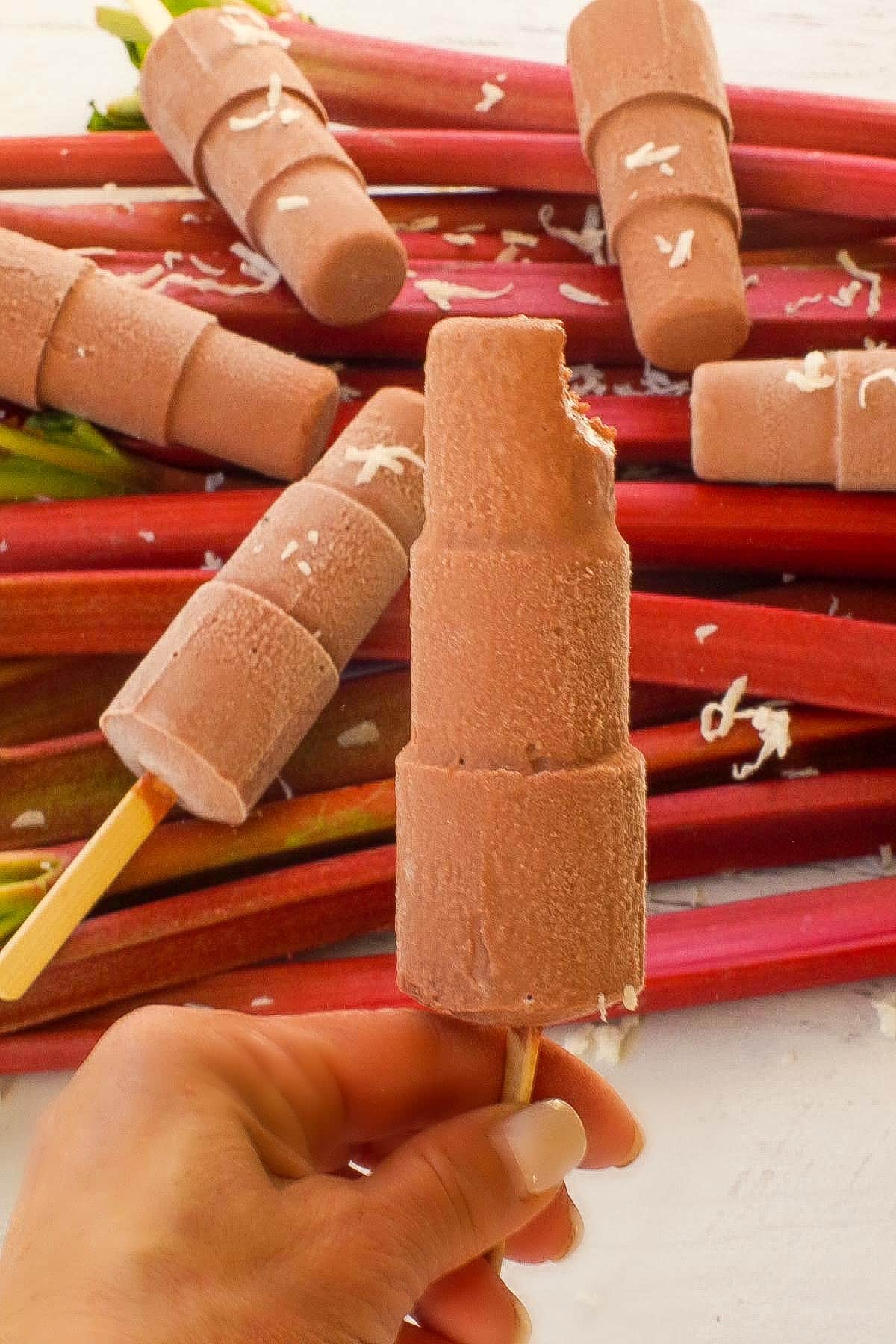 Chocolate Rhubarb Popsicle (with a bite taken out at the top) being held up in front of a pile of rhubarb with more popsicles on top