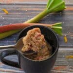Rubarb Mug Muffin piece being held up on a spoon over a mug, with 2 rhubarb stalks in the background.