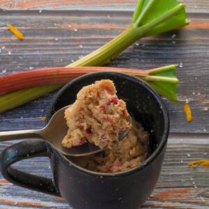 Rubarb Mug Muffin piece being held up on a spoon over a mug, with 2 rhubarb stalks in the background.