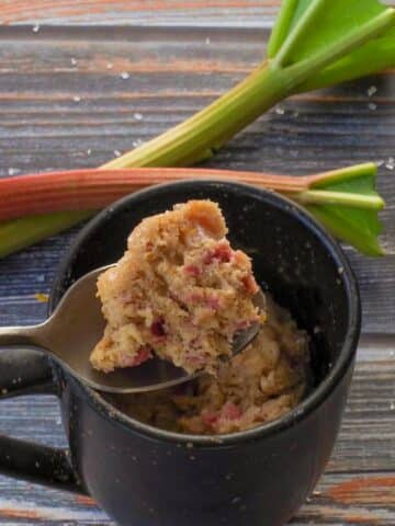 Rubarb Mug Muffin piece being held up on a spoon over a mug, with 2 rhubarb stalks in the background.