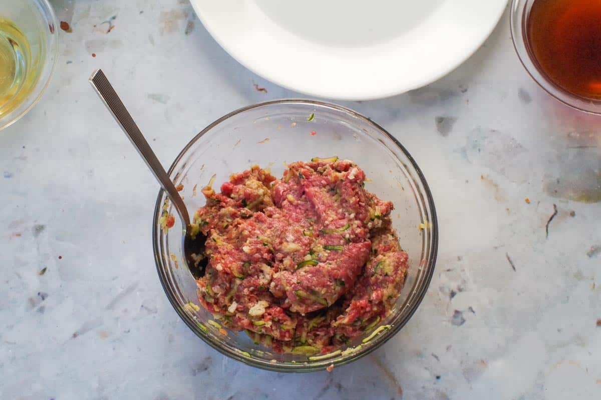 Ground beef, zucchini, spices,egg and breadcrumbs in a glass bowl mixed together.