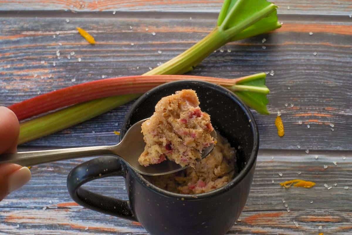 Rubarb Mug Muffin piece being held up on a spoon over a mug, with 2 rhubarb stalks in the background.