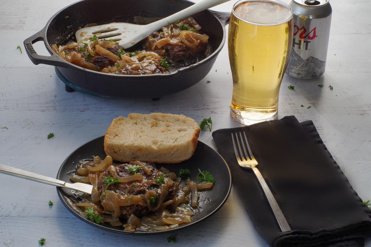 Healthy Salisbury Steak on a black plate with a piece of Beer Cheese Bread and glass of beer and a pan of more Salisbury Steak in the background.