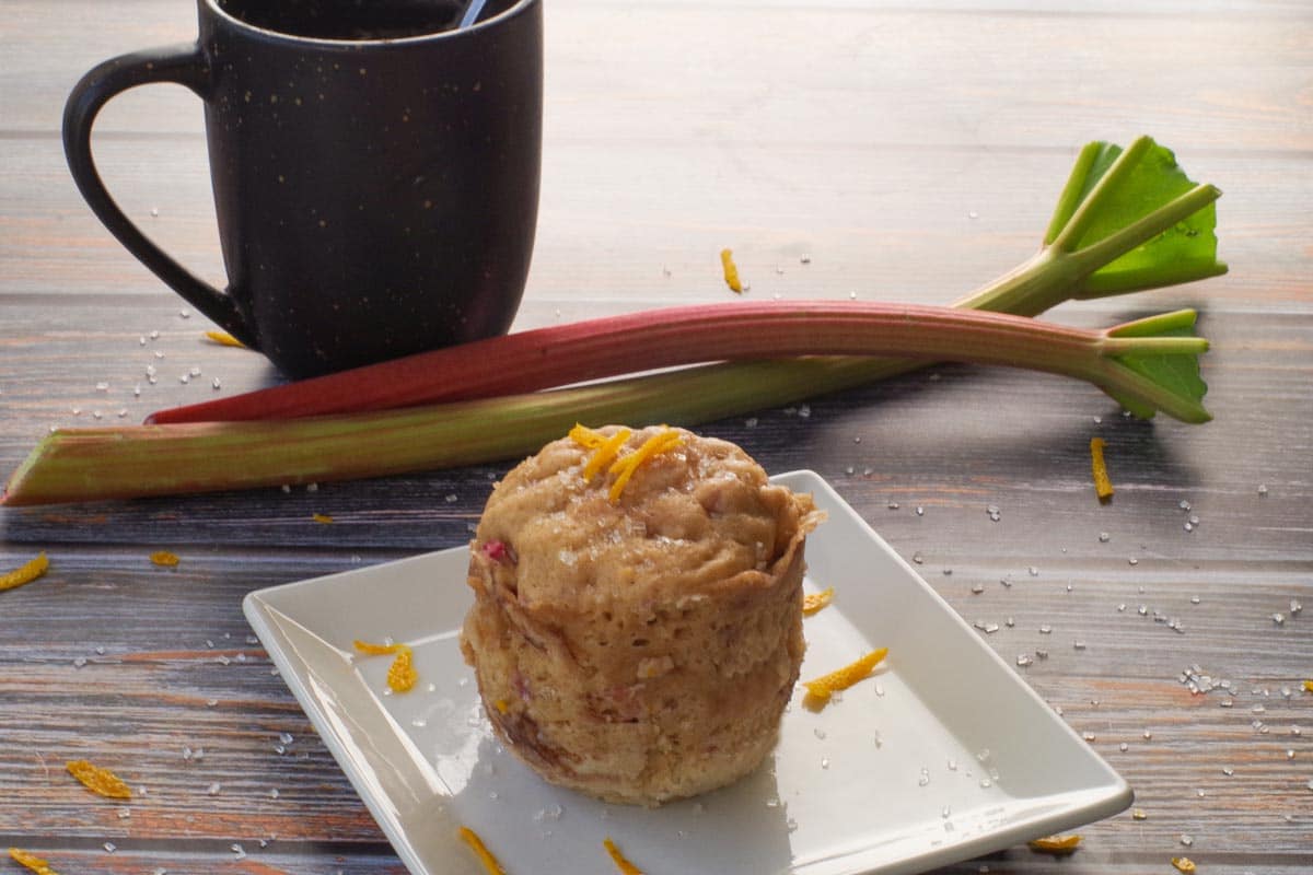Whole Rhubarb Microwave Muffin (in a Mug) on a white square plate with a mug and some rhubarb in the background.