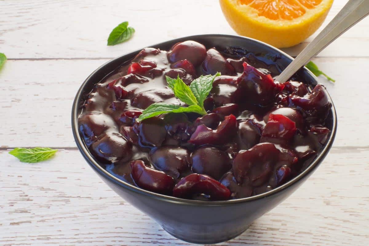 Fresh Cherry sauce in a black bowl with a spoon and a sprig of mint on top and ½ and orange in the background.