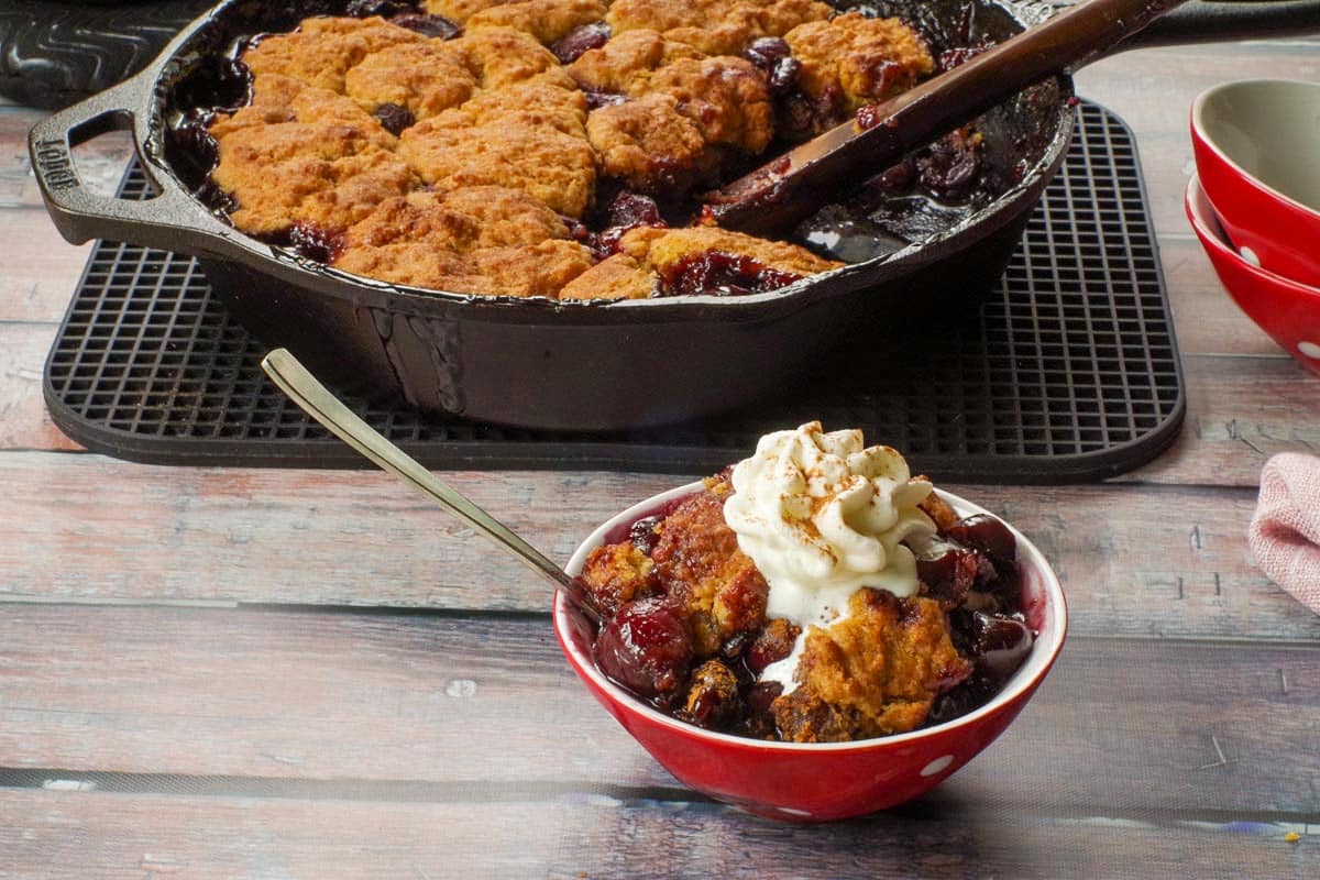 Cherry cobbler in a bowl with whipped cream with more fresh cherry cobbler in a skillet in the background.