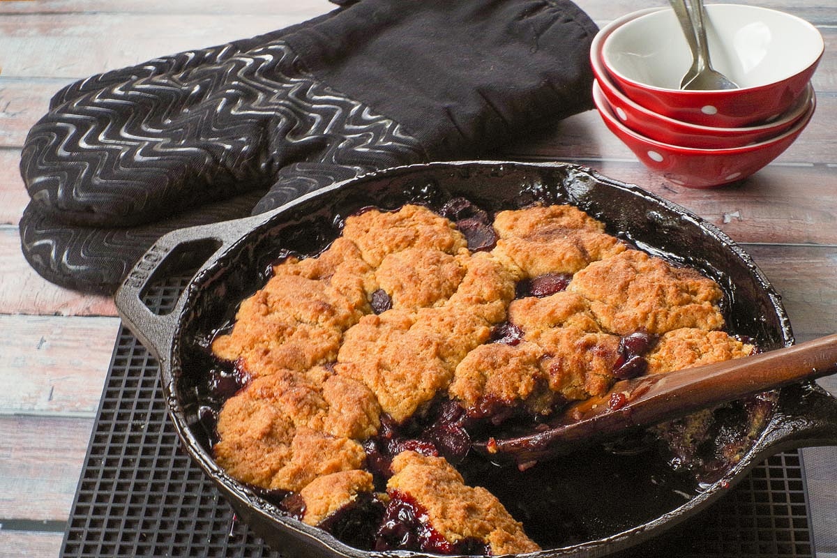 Fresh Cherry Cobbler in a cast iron pan with a wooden spoon in a spot where a serving of fresh cherry cobbler was scooped out.