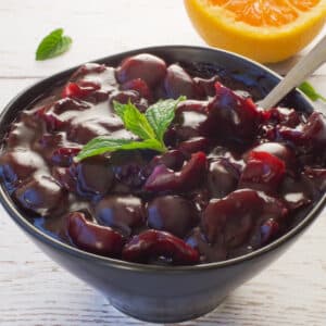 Fresh Cherry sauce in a black bowl with a spoon and a sprig of mint on top and ½ and orange in the background.