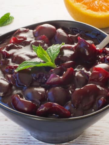 Fresh Cherry sauce in a black bowl with a spoon and a sprig of mint on top and ½ and orange in the background.