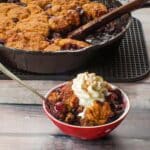 Cherry cobbler in a bowl with whipped cream with more fresh cherry cobbler in a skillet in the background.
