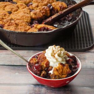 Cherry cobbler in a bowl with whipped cream with more fresh cherry cobbler in a skillet in the background.