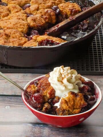 Cherry cobbler in a bowl with whipped cream with more fresh cherry cobbler in a skillet in the background.