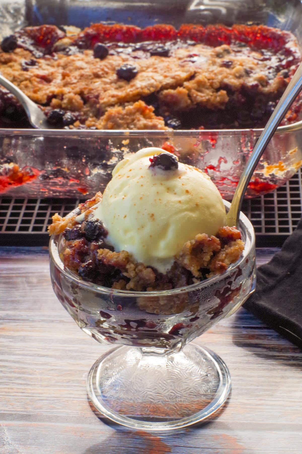 Old- Fashioned Saskatoon berry crumble in a glass dessert dish in front of more Saskatoon berry crumble in a square glass baking pan in the background.