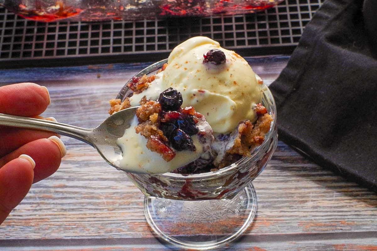 Spoonful of Saskatoon berry crumble with ice cream in front of a dish with more of the same.