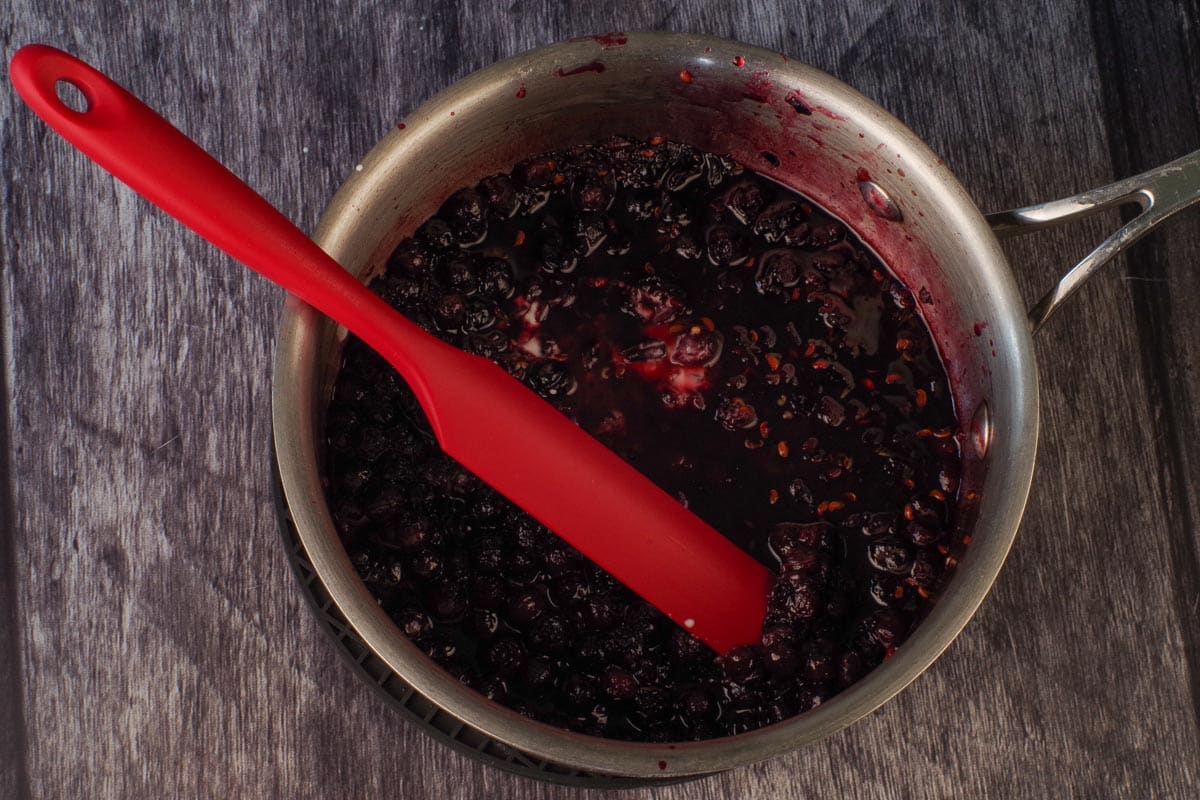 Cornstarch slurry and almond extract added to Saskatoon Berry mixture in small saucepan with red jar spatula.