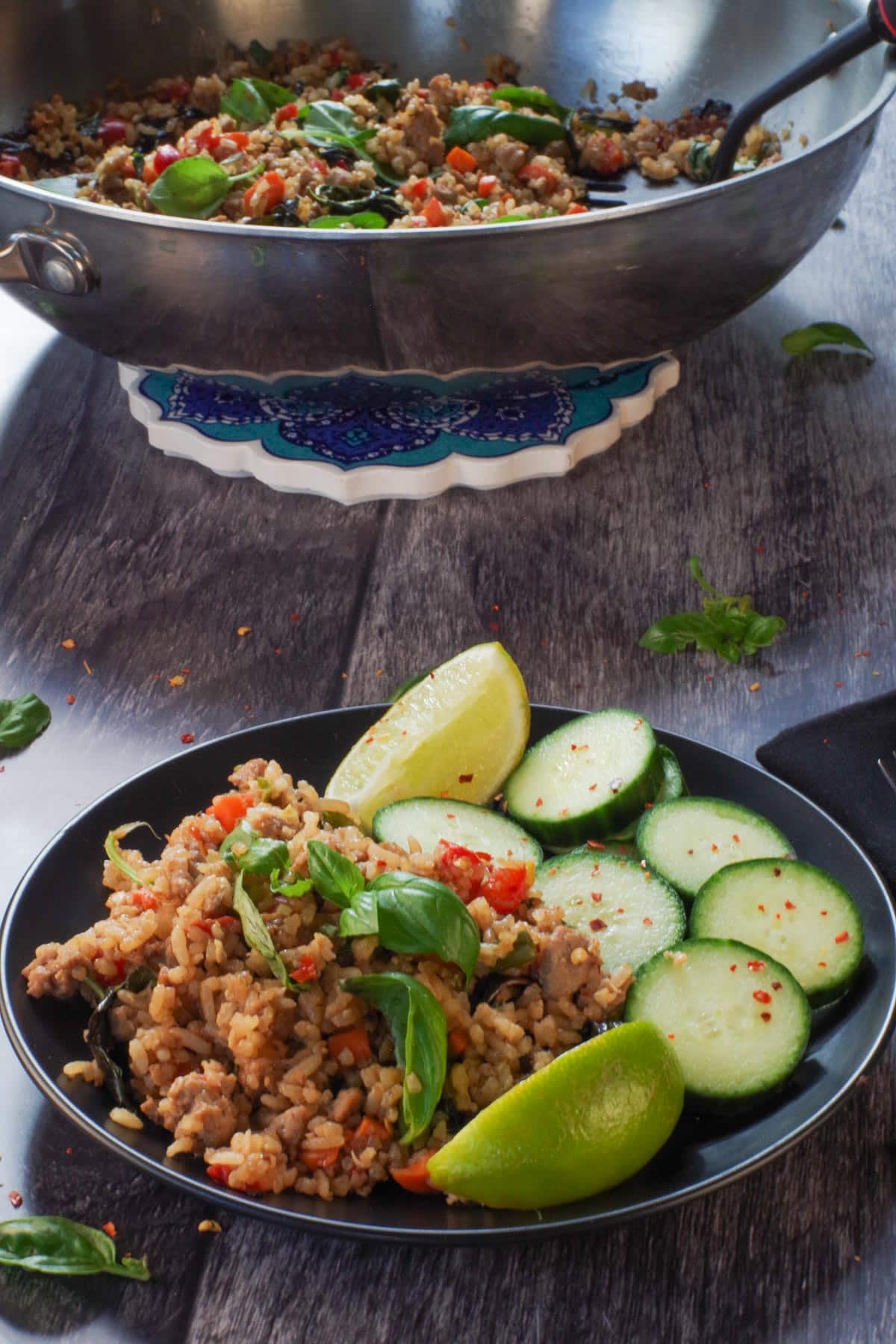 Thai Basil Fried Rice with chicken on a black plate with cucumbers and more thai basil fried rice in a wok in the background.