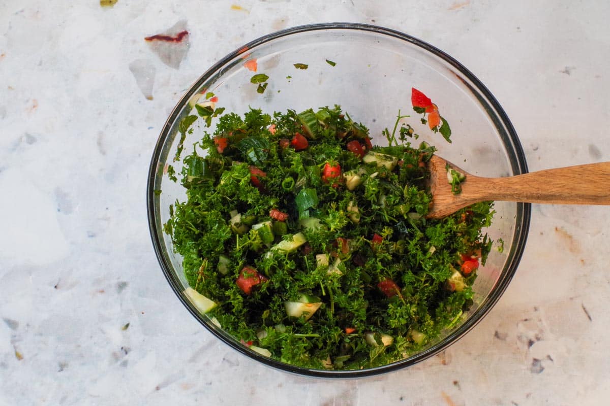 Parsley, onions, tomatoes and cucumbers mixed together in large glass bowl. 