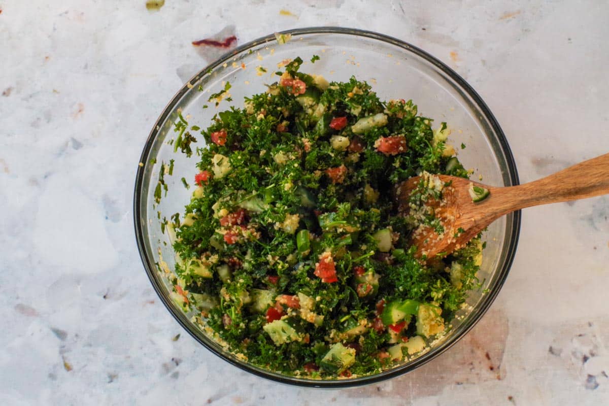 Lemon soaked bulgur mixture added to the greens in a large glass bowl (to make Tabouli salad) with a wooden spoon.