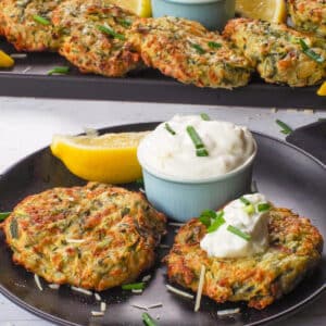 2 Mediterranean Air Fryer Zucchini Fritters on a black plate with tzatziki and a tray of more in the background