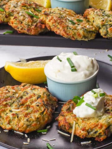 2 Mediterranean Air Fryer Zucchini Fritters on a black plate with tzatziki and a tray of more in the background