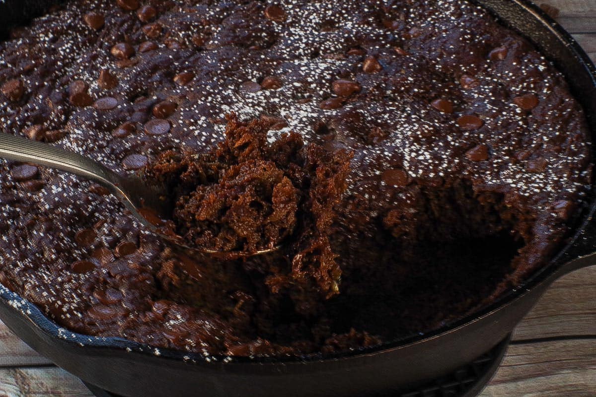Chocolate Zucchini Spoon Cake being held up on a spoon over a skillet of more cake.