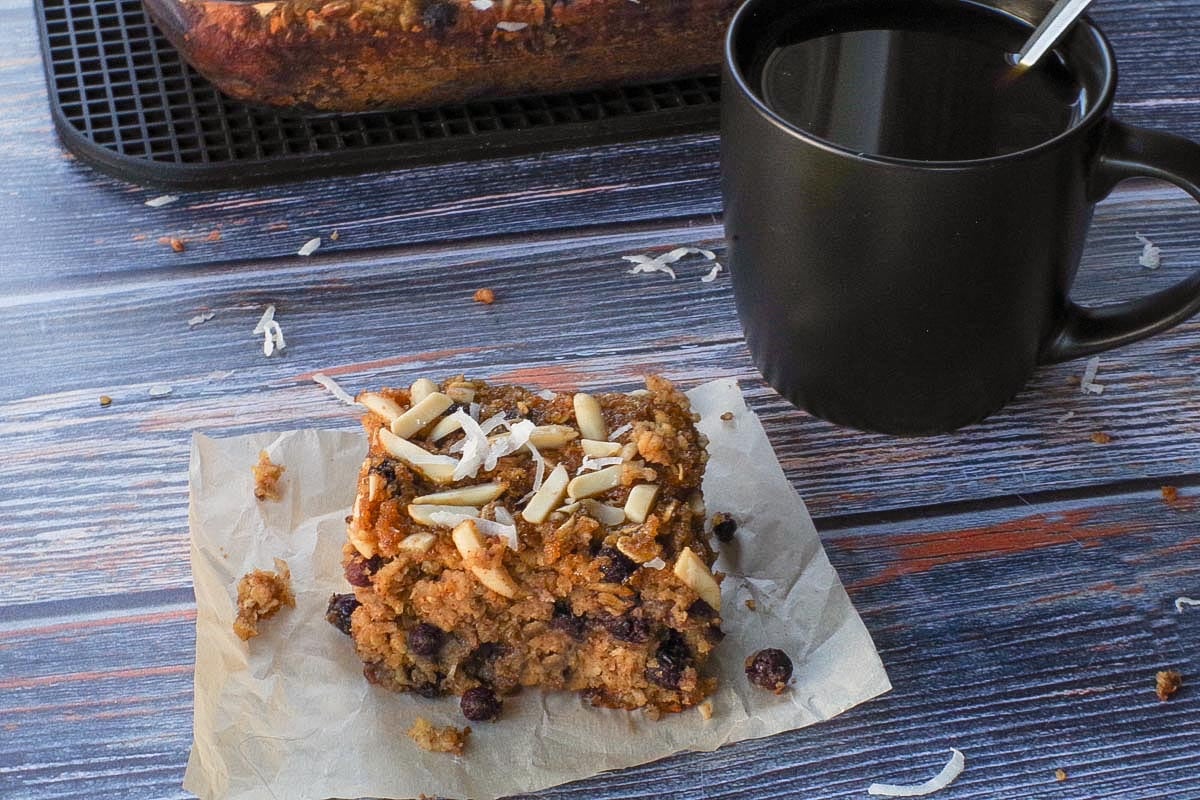 A Saskatoon Berry Oatmeal Square on a piece of parchment paper with a cup of coffee and more Saskatoon Berry Oatmeal Bar in a glass pan in the background.