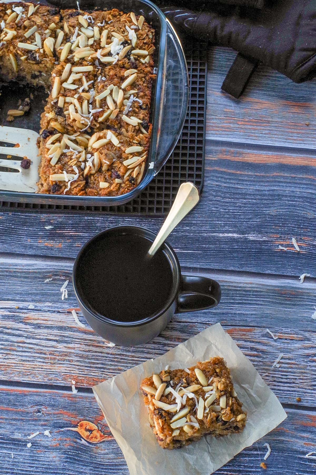 Photographed from above: A Saskatoon Berry Oatmeal Square on a piece of parchment paper with a cup of coffee and more Saskatoon Berry Oatmeal Bar in a glass pan in the background.