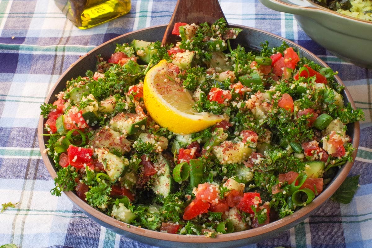 A bowl of Mediterranean Tabouleh Salad with a dark wooden spoon in it, on a blue and white checked table cloth.