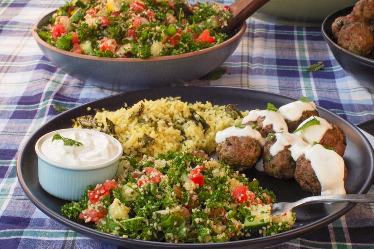 Mediterranean Tabouli on a black plate with Greek meatballs and Lazy Dolmades, with a bowl of more Tabouli in the background.