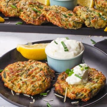 2 Mediterranean Air Fryer Zucchini Fritters on a black plate with tzatziki and a tray of more in the background
