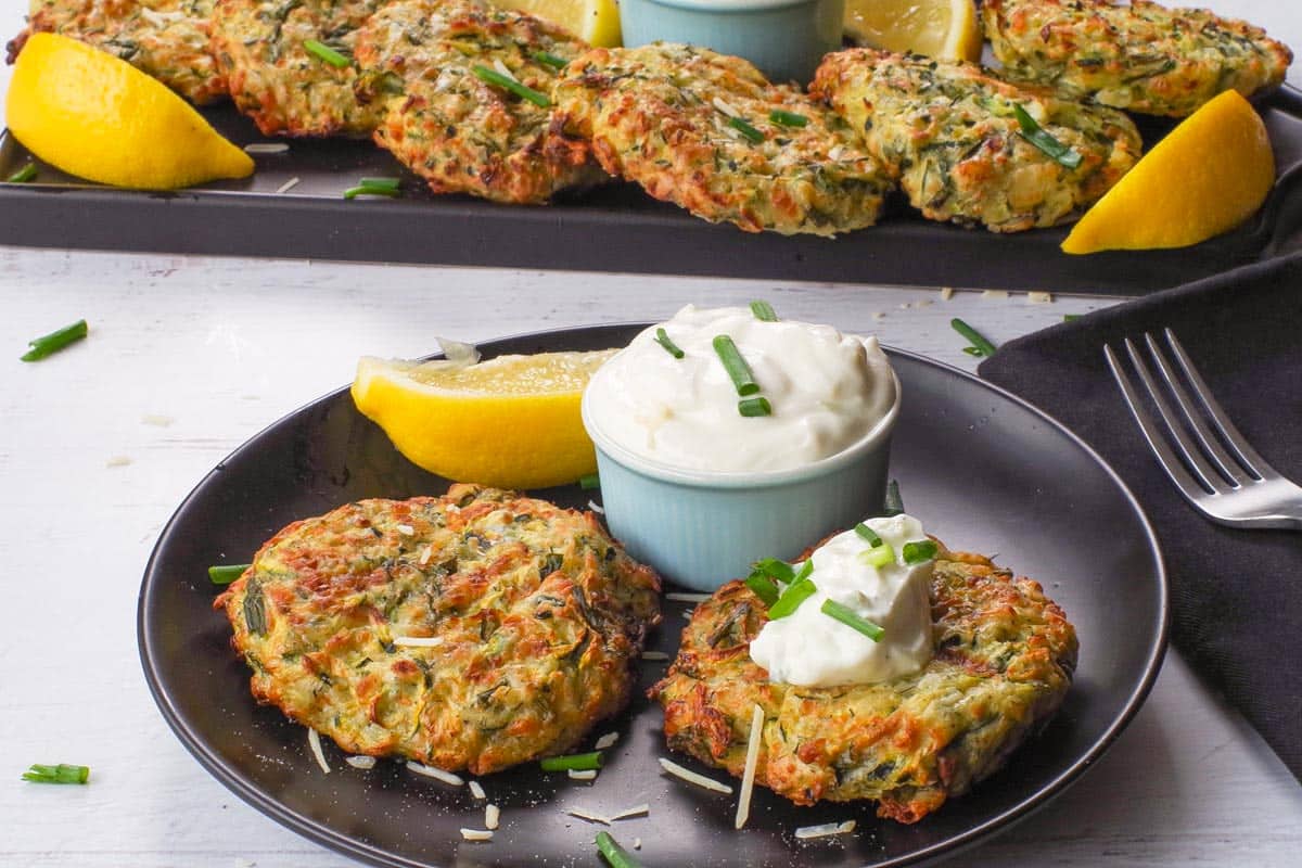 2 Mediterranean Air Fryer Zucchini Fritters on a black plate with tzatziki and a tray of more in the background
