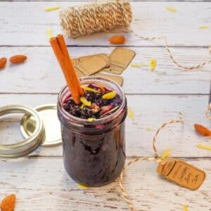 Saskatoon Berry Pie Jam in a glass jam jar with a cinnamon stick in the jar and lemon zest, surrounded by labels, and string.