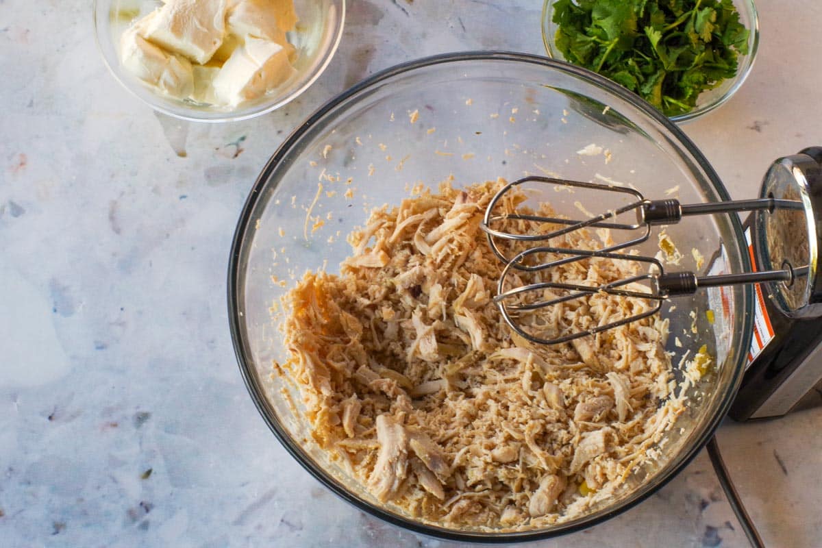 Chicken being shredded in a large glass bowl with a hand mixer.