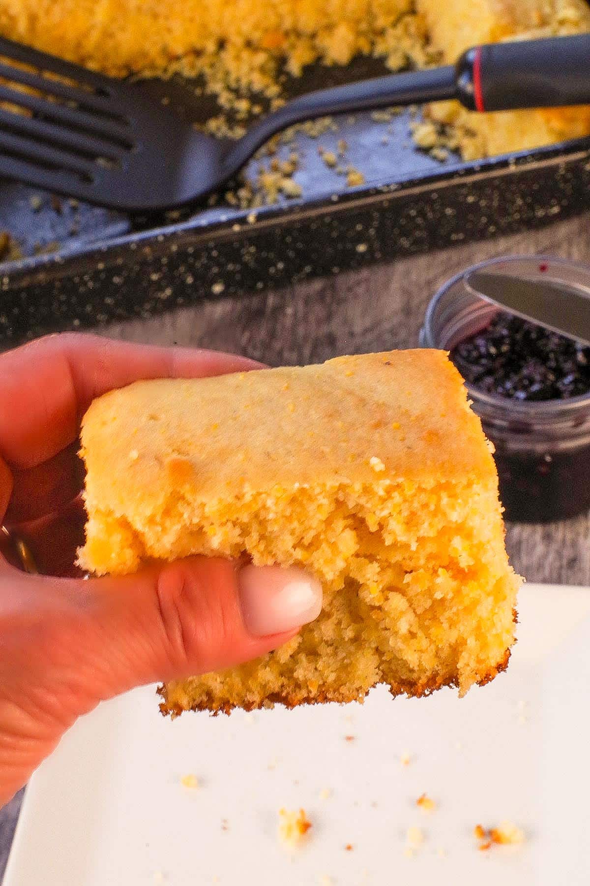 A piece of Old Fashioned Country Cornbread being held up over a plate with a pan with more cornbread in the background.