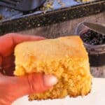 A piece of Old Fashioned Country Cornbread being held up over a plate with a pan with more cornbread in the background.