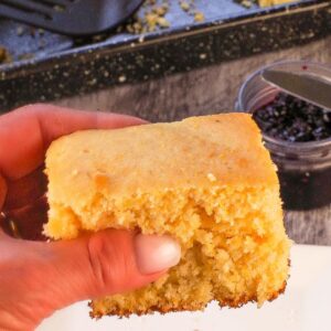 A piece of Old Fashioned Country Cornbread being held up over a plate with a pan with more cornbread in the background.
