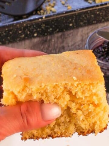 A piece of Old Fashioned Country Cornbread being held up over a plate with a pan with more cornbread in the background.